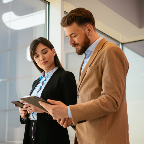 Two business professionals, a woman and a man, stand near a large window. They are both wearing business attire; the woman has on a dark-colored blazer and the man has a light beige or tan blazer. Both appear to be looking at and holding digital tablets or clipboards. The woman is looking down at the tablet or clipboard in her hands, and the man is looking at the tablet/clipboard in front of him while glancing over at the woman. The building exterior, visible through the window, reflects a light blue sky. The background suggests an interior office space.