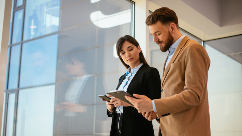 Two business professionals, a woman and a man, stand near a large window. They are both wearing business attire; the woman has on a dark-colored blazer and the man has a light beige or tan blazer. Both appear to be looking at and holding digital tablets or clipboards. The woman is looking down at the tablet or clipboard in her hands, and the man is looking at the tablet/clipboard in front of him while glancing over at the woman. The building exterior, visible through the window, reflects a light blue sky. The background suggests an interior office space.